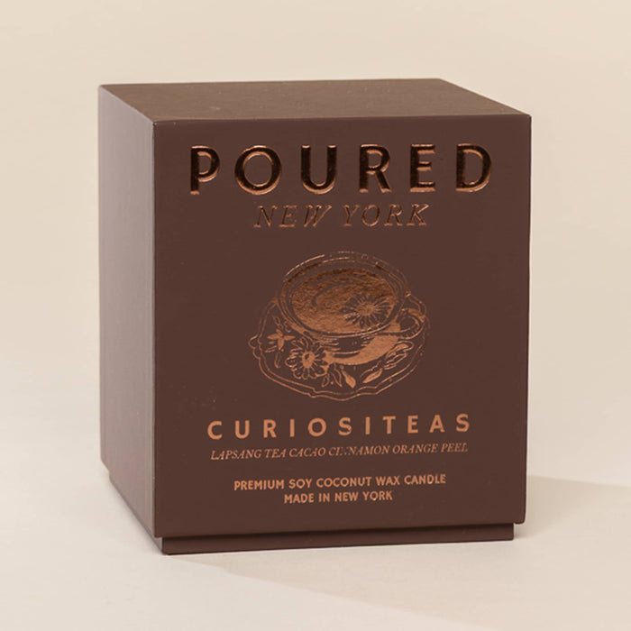 A square dark brown cardboard box for a Poured New York Curiositeas candle, featuring embossed metallic brown lettering and an illustration of a teacup on a light beige background.