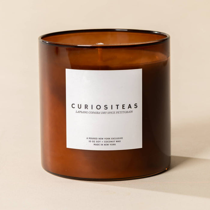 An amber glass jar candle with a white rectangular label reading 'CURIOSITEAS' and smaller text below, photographed on a warm beige background.