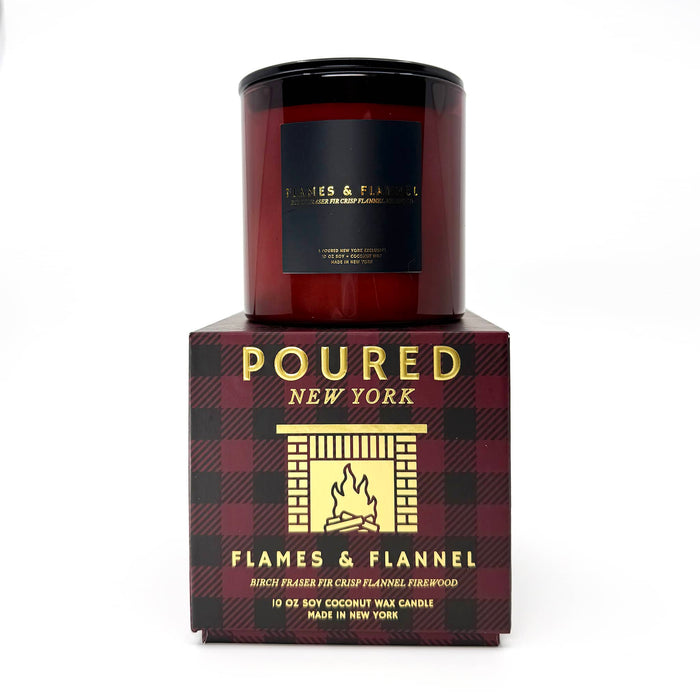A candle in a dark red glass vessel sits on top of its retail box. The box is dark red and black plaid with gold lettering that reads 'POURED NEW YORK' and 'FLAMES & FLANNEL'.