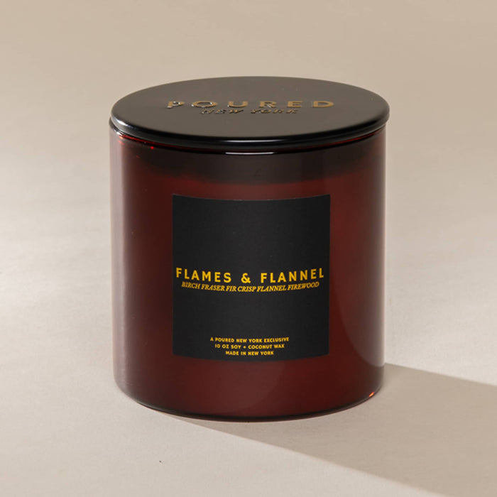 An amber glass jar candle with a black lid and a black label featuring gold text that reads 'POURED BY NEW YORK and 'FLAMES & FLANNEL' on a beige background.