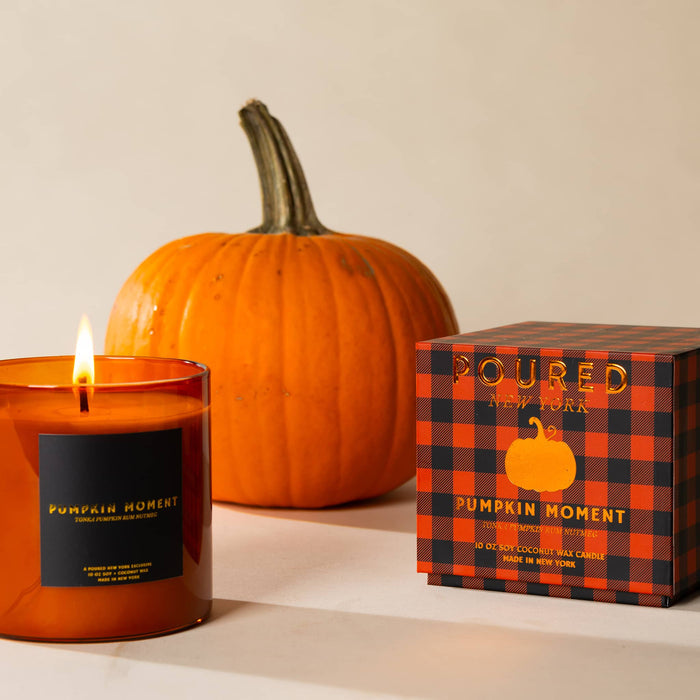 A lit amber glass candle labeled 'Pumpkin Moment' sits beside an orange-and-black plaid product box with the same name, with a whole pumpkin in the background on a warm beige surface.
