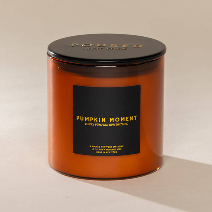 An amber glass jar candle with a black metal lid and a black label reading 'PUMPKIN MOMENT' in gold lettering, photographed on a warm beige background.