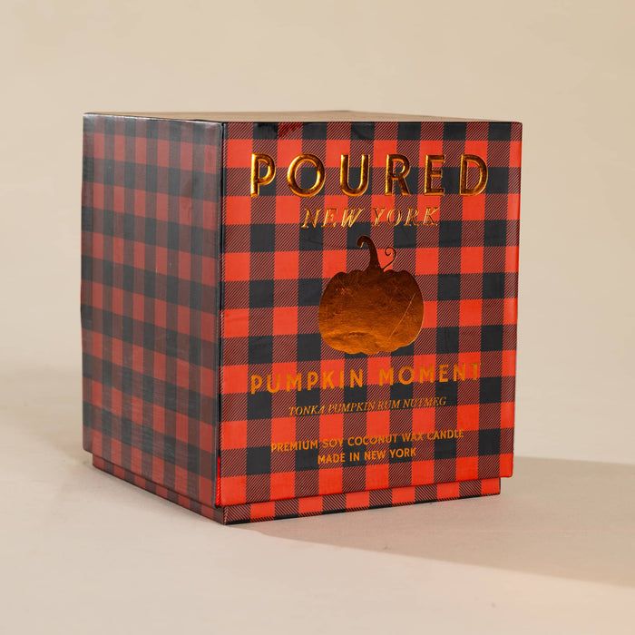 A square cardboard box for a candle, featuring an orange and black buffalo plaid pattern with gold foil text reading 'POURED NEW YORK' and 'PUMPKIN MOMENT.'