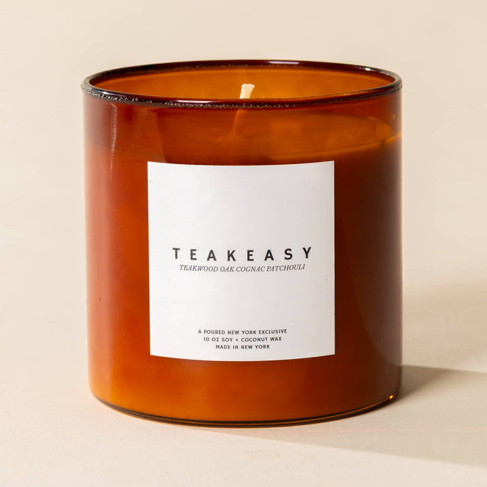 An amber glass jar candle with a white rectangular label reading TEAKEASY, photographed on a warm beige background.