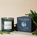 A lit green glass candle next to its dark green box, both labeled 'POURED NEW YORK, UPSTATE HIBERNATE', styled with green pine branches on a beige background.