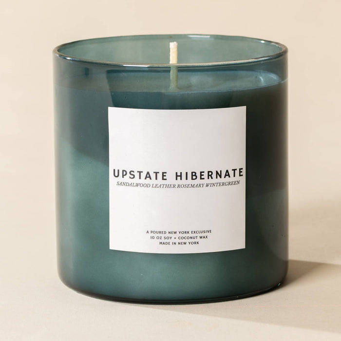 A dark green glass jar candle with a white label reading 'UPSTATE HIBERNATE' and a single white wick, photographed on a warm beige background.