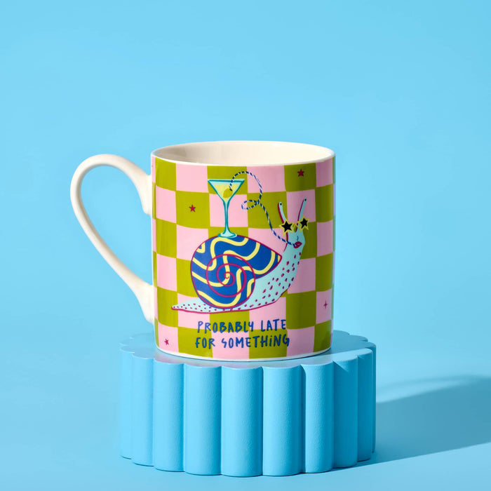 A white ceramic mug featuring a colorful design with a pink and lime green checkered pattern, a cartoon snail with a cocktail glass on its shell, and the text 'PROBABLY LATE FOR SOMETHING'. Handle is on the left and it's displayed on a blue fluted pedestal against a solid blue background.