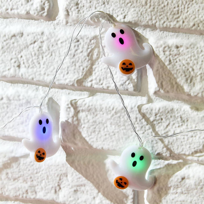 Three white ghost-shaped string lights glowing pink, blue, and green, each holding an orange jack-o'-lantern, hanging against a white brick wall.
