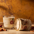 Two scented candles in clear glass jars with gold rims, one standing upright and one lying on its side, displayed on a wooden surface with dried citrus slices and cinnamon sticks against a warm textured background. The label reads 'CRUSH'.