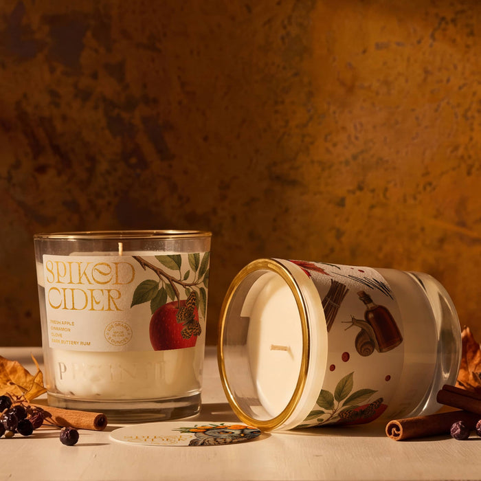 Two glass candles with white wax and a printed label featuring a red apple and warm spices, styled on a tabletop with cinnamon sticks and dried leaves against a textured warm brown background. One candle stands upright and the other lies on its side showing the lid.