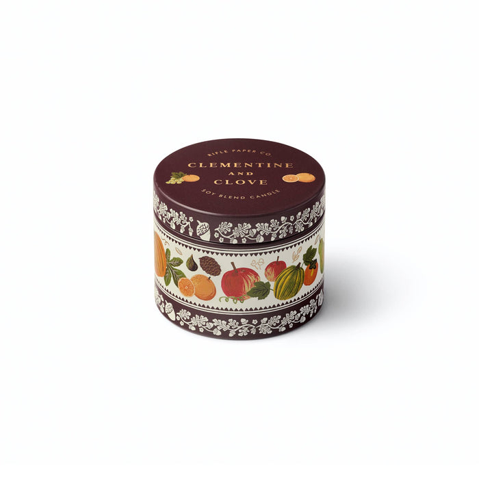 A round metal tin candle labeled 'CLEMENTINE AND CLOVE' by Rifle Paper Co., featuring a dark brown lid and a cream-colored band decorated with illustrations of citrus fruits and autumn leaves.