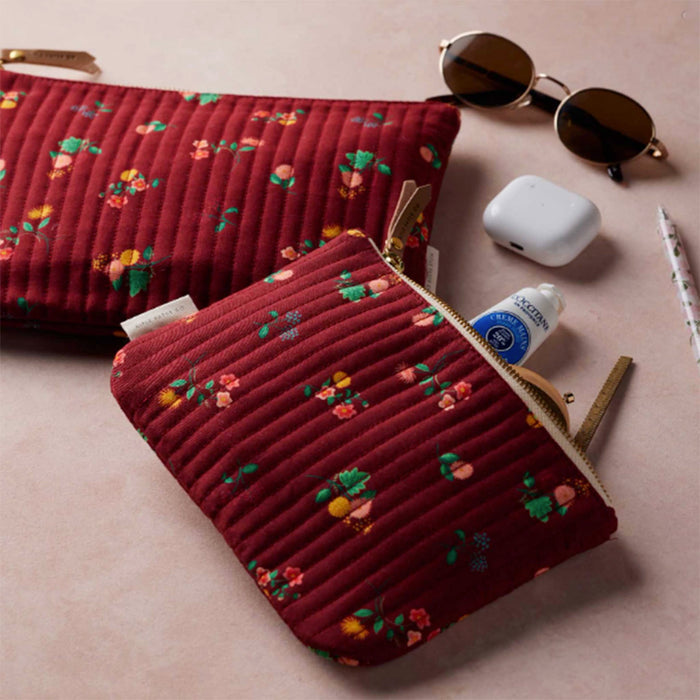 A pair of quilted burgundy zip pouches with a small multicolor floral print, styled with sunglasses, a white earbud case, and a tube of lip balm on a light pink background.