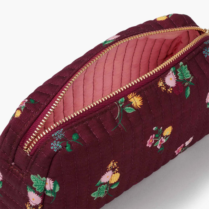 A close-up of a quilted burgundy cosmetic pouch with a floral print, shown open to reveal a pink interior and gold zipper.