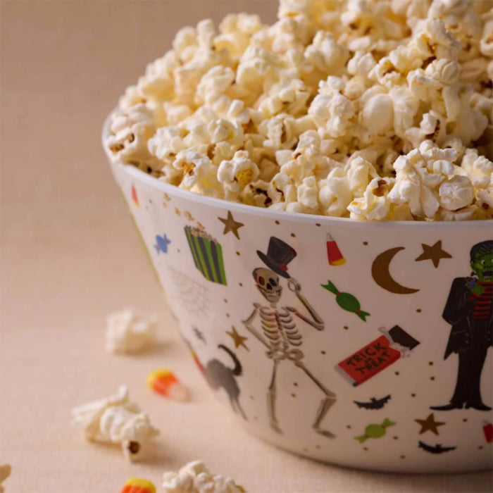 A white melamine bowl decorated with Halloween-themed illustrations, filled with fluffy popcorn on a warm beige surface.