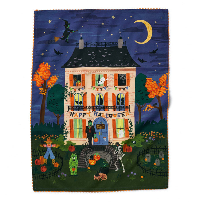 A rectangular tea towel with a Halloween-theme featuring a whimsical illustration of a haunted house at night, with pumpkins, bats, a crescent moon, stars, and characters in front of the door. The design has a dark navy sky, orange trees, and a cream house, with an orange ric-rac border along the edges.