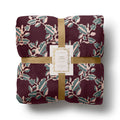 A folded fleece blanket with a botanical leaf pattern in burgundy, sage green, and blush pink, wrapped with a gold ribbon and a white paper label that reads 'Rifle Paper Co FLEECE BLANKET'.