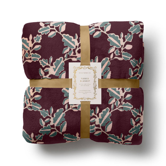 A folded fleece blanket with a botanical leaf pattern in burgundy, sage green, and blush pink, wrapped with a gold ribbon and a white paper label that reads 'Rifle Paper Co FLEECE BLANKET'.
