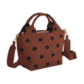 A brown handbag with black polka dots, featuring dual top handles, a top zipper, and an adjustable crossbody strap with gold-tone hardware, front angle view, shown on a plain white background.