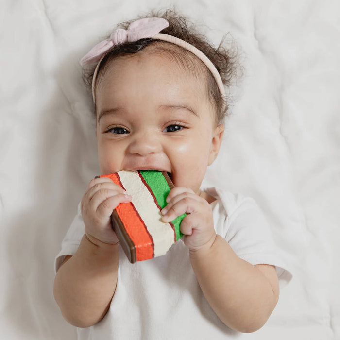 Baby holding a teether that looks like a rainbow cookie, on a white background.