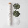 Glass tube with cork stopper filled with bath salt soak with piles of ingredients beside it, on a white background.