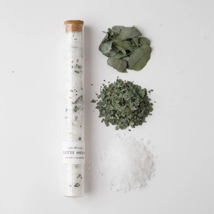 Glass tube with cork stopper filled with bath salt soak with piles of ingredients beside it, on a white background.