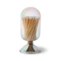 A clear glass cloche with an iridescent finish lid containing wooden safety matches, with a small gold triangular brand sticker on the base, photographed on a white background.