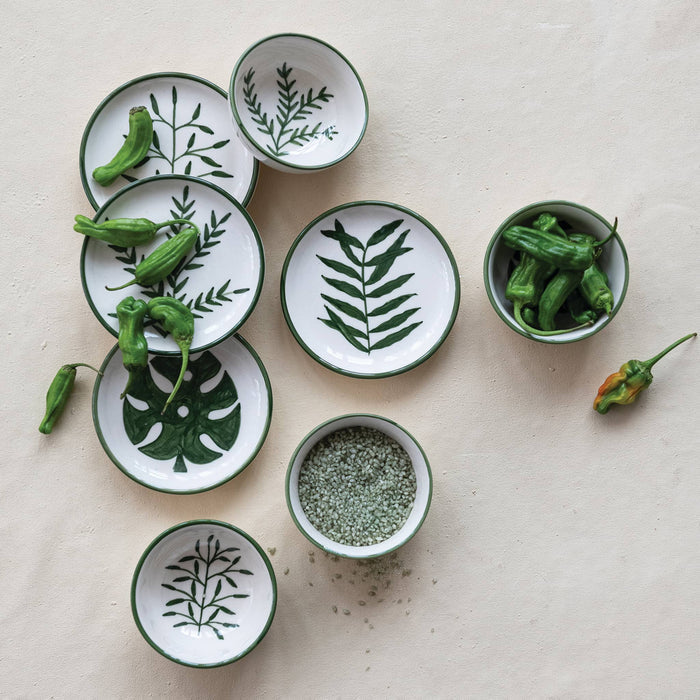A set of small cream ceramic dishes and bowls with green botanical leaf patterns, arranged on a light beige surface with green peppers and coarse sea salt.