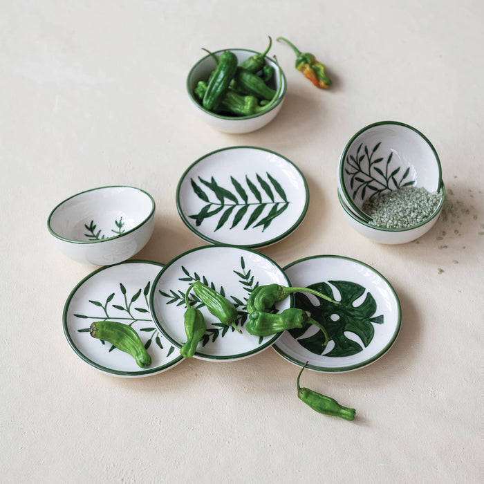 A set of white small ceramic bowls and dishes with green leaf patterns, styled with green peppers and a small bowl of herbs on a light beige textured background.