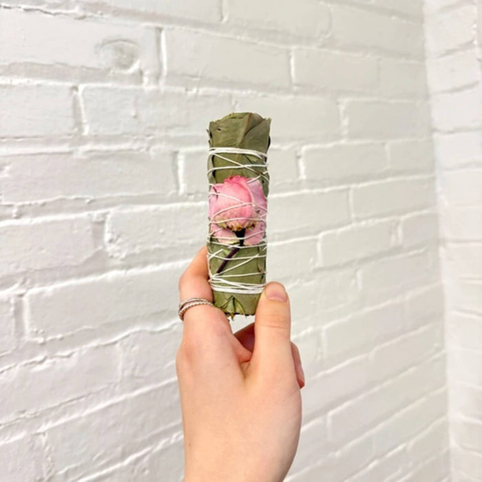 Hand holding a dried eucalyptus smudge bundle with a dried pink rose, tied up with cotton string, against a white brick wall.