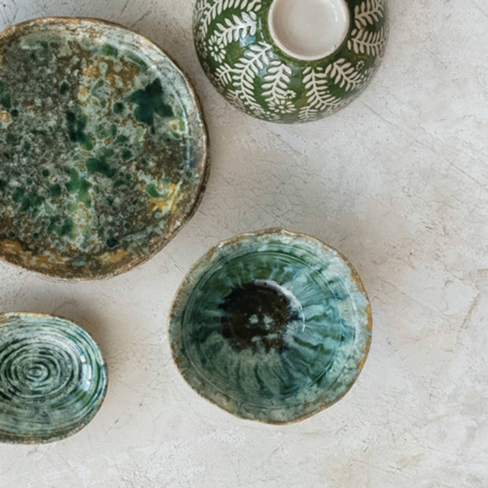A set of handmade ceramic tableware in earthy green and brown glazes, including a large plate, a patterned bowl, and two small bowls, arranged on a textured light beige surface.