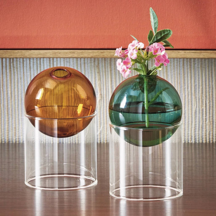 Two decorative glass vases displayed on a wooden surface against a striped wall and terracotta background. Two translucent glass vases with spherical tops in amber and green, each resting on a clear cylindrical base. The green vase has a sprig of pink flowers and is filled with water.