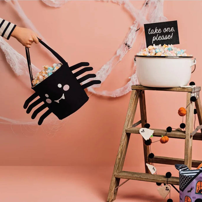 A Halloween-themed black spider bucket bag filled with wrapped candy, held by a child's hand against a pink backdrop. To the right, a rustic wooden ladder holds a white enamel tub of candy with a 'take one, please!' sign.