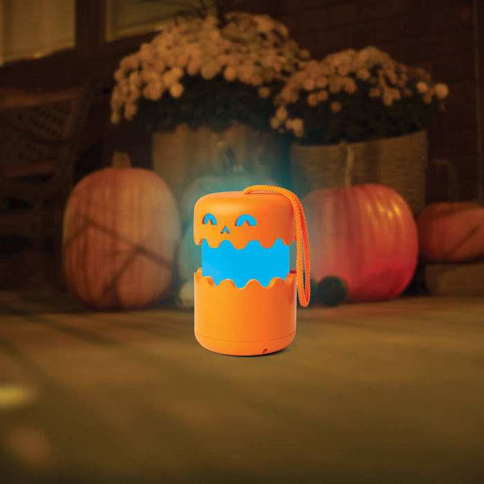 An orange Halloween-themed pop-up lantern with a glowing blue mouth and jack-o'-lantern face, placed on a wooden surface with pumpkins and potted plants in the background.