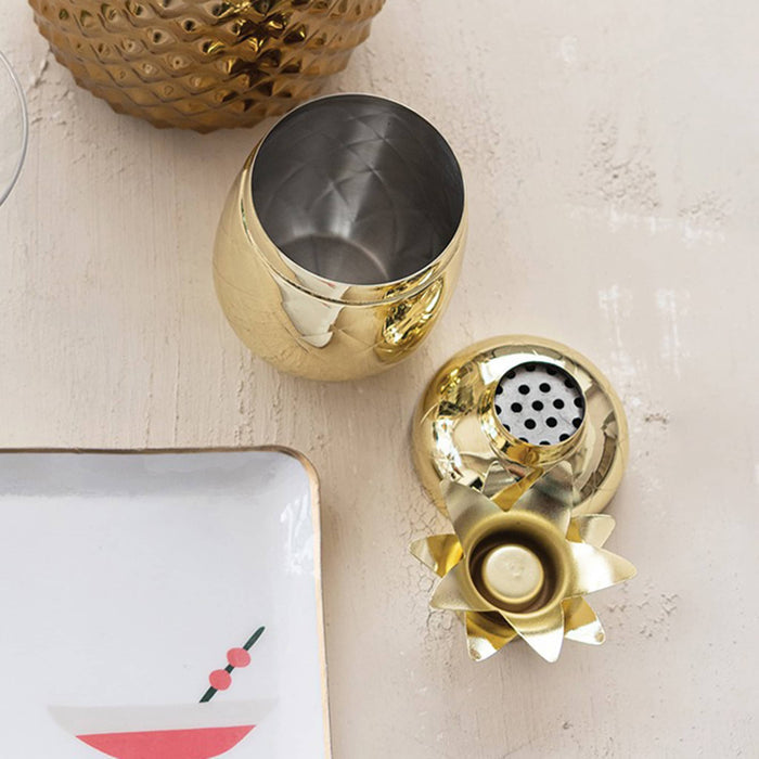 Gold and stainless steel pineapple cocktail shaker, lid off, on a light surface with a decorative item in the background.