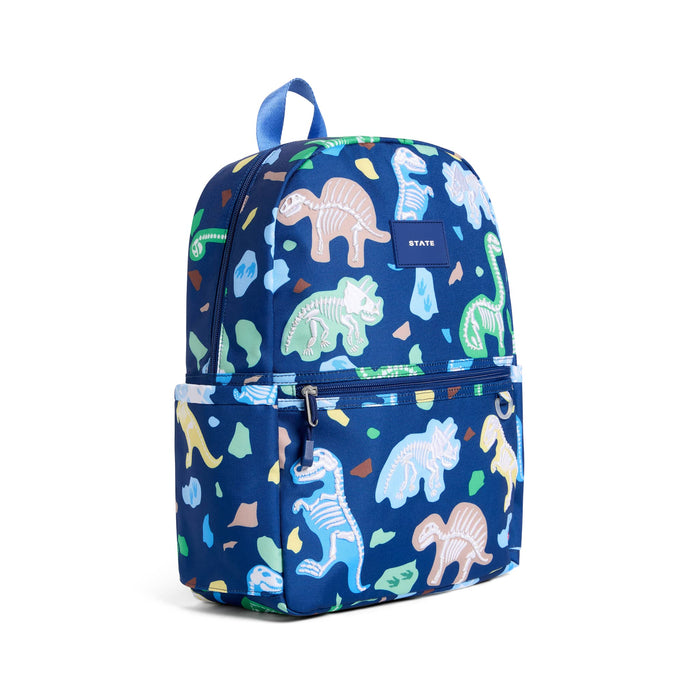 A blue backpack with an all-over dinosaur skeleton and stone pattern, featuring a top handle, front zippered pocket, and a small 'STATE' brand label on the upper right side. Front angle view on a plain white background.