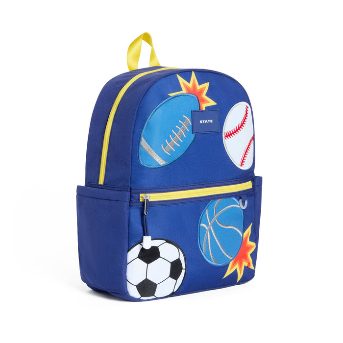 A royal blue backpack decorated with sports ball graphics including a football, baseball, soccer ball, basketball, and starburst shapes, with yellow zipper accents and a yellow top handle. Front angle view on a plain white background.