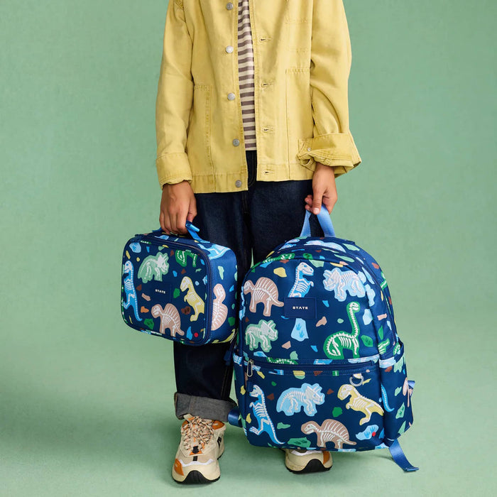 Child holding a blue backpack and lunch bag with dinosaur pattern on a green background.