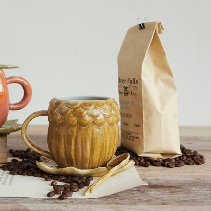 An acorn-shaped ceramic mug  sits on a matching saucer on a wooden table, surrounded by coffee beans and a paper bag of coffee from River Falls Coffee Co.