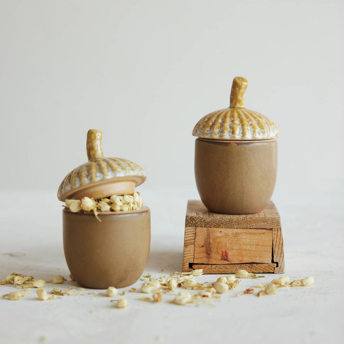 Two small acorn-shaped ceramic containers with matching lids, one open with dried flowers inside and one closed, displayed on a light background with one container resting on a wooden block.