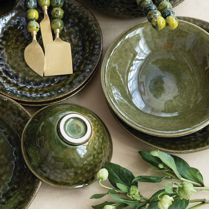 A styled flat-lay of green glazed ceramic tableware, including bowls, plates, and a small covered dish, arranged on a light neutral surface with decorative green-beaded utensils and leafy stems.