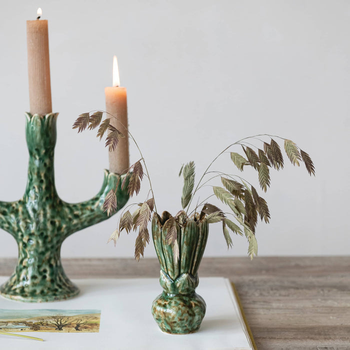A green glazed ceramic vase holding dried ornamental grass stems, styled on a tabletop beside a matching green candlestick with lit candles against a soft light gray background.