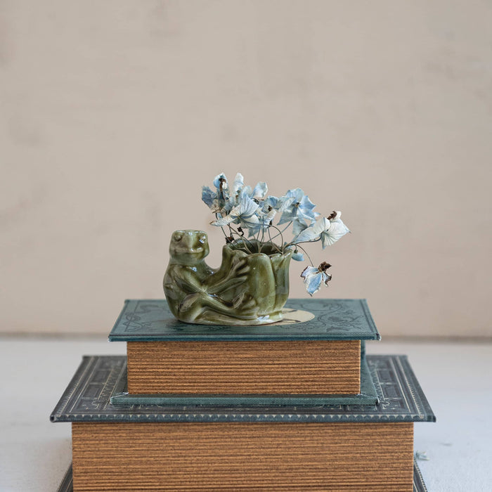 A small green glazed stoneware figurine shaped like a seated frog holding a lotus flower vase with small blue and white flowers, on top of a stack of books against a plain beige background.