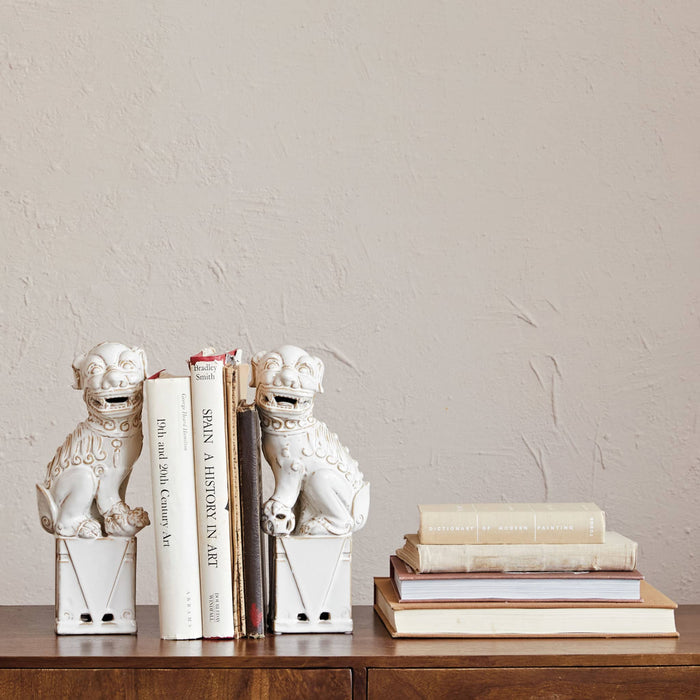 Two white ceramic lion statues on a wooden surface with books stacked nearby against a beige wall.