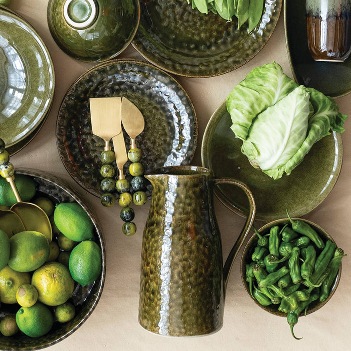 A styled overhead flat-lay of olive green ceramic tableware, including a pitcher, plates, bowls, and serving utensils, arranged with limes, cabbage, and green peppers on a neutral beige surface.