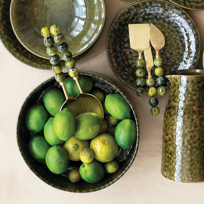 A styled flat-lay of rustic ceramic tableware on a light beige surface, featuring a hammered green bowl filled with limes and gold-toned serving utensils with bead handles, alongside matching plates and a textured green pitcher.