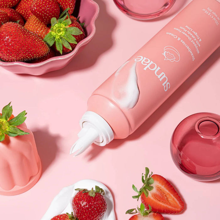 Sundae Strawberries and Cream scented whipped shower foam in pink bottle, cap off, with strawberries and whipped cream on a pink background.