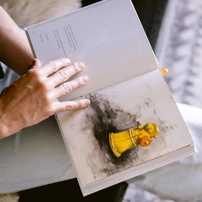 An open book with an art print being held by a person's hand, showing a yellow fire hydrant illustration on the right page and a poem on the left.
