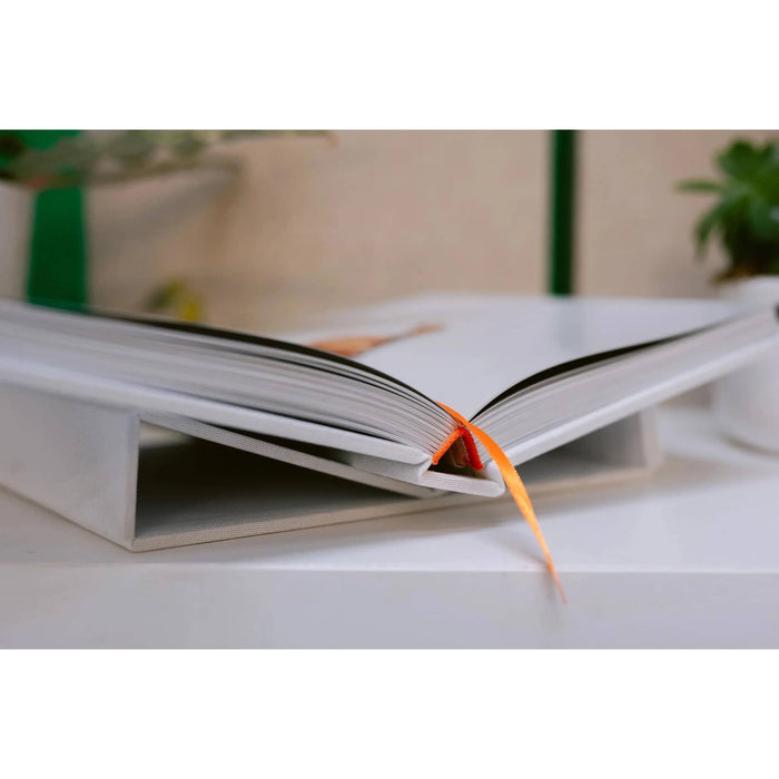 A white book stand holding an open book, with a bright orange ribbon bookmark hanging down, set on a white desk with a softly blurred indoor background.