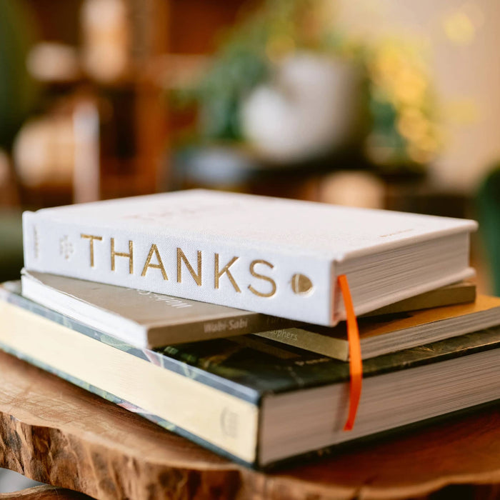 A stack of hardcover books on a rustic wooden slice table, with the top book titled 'THANKS' in gold lettering and an orange ribbon bookmark visible.
