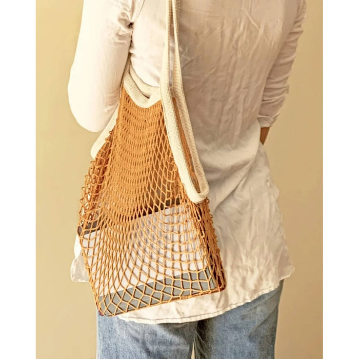A person wearing a white long-sleeve shirt and blue jeans carries a yellow cotton net shoulder bag with a thick cream strap against a plain beige background.